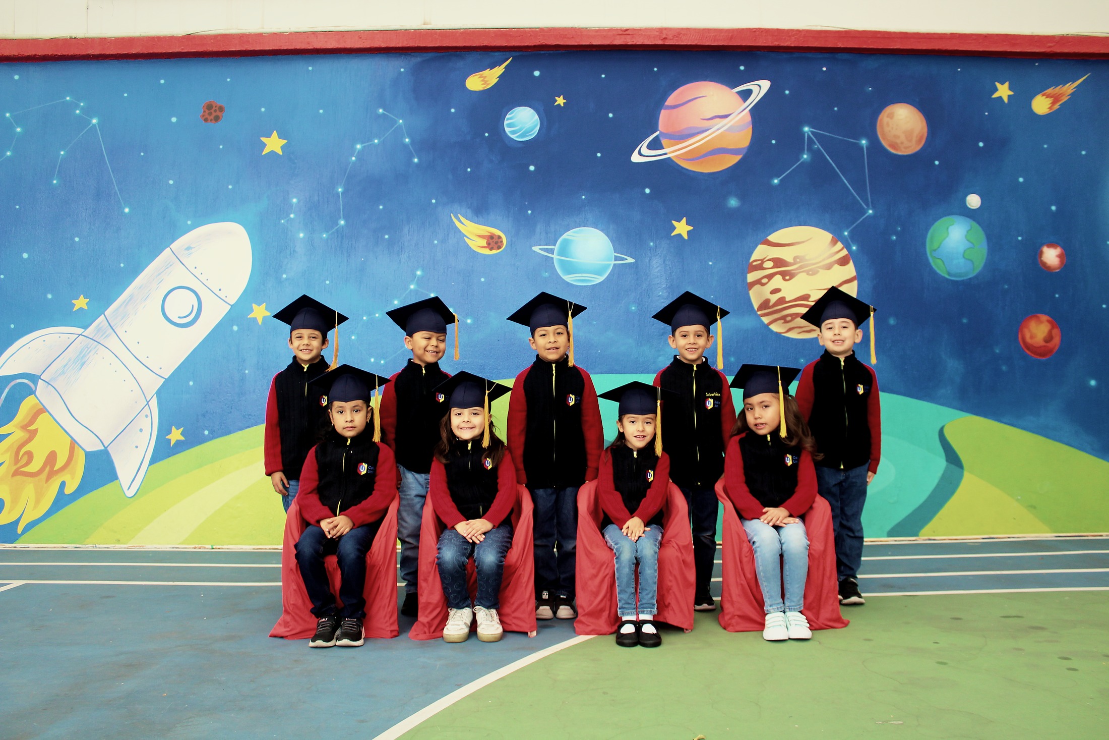 Generación 2026 de Kínder con toga y birrete frente al mural del espacio